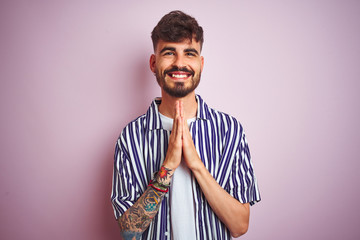 Wall Mural - Young man with tattoo wearing striped shirt standing over isolated pink background praying with hands together asking for forgiveness smiling confident.