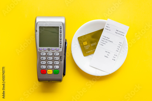 Electronic payments in restaurant. Bank card near aquiring terminal and bill on yellow background top view