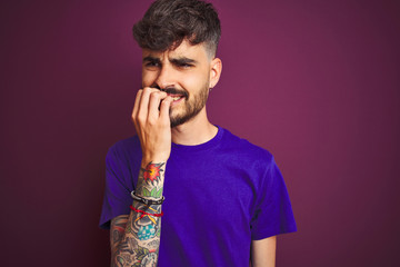 Sticker - Young man with tattoo wearing t-shirt standing over isolated purple background looking stressed and nervous with hands on mouth biting nails. Anxiety problem.