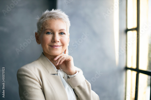Fotomural Elegant middle aged businesswoman with stylish pixie hair standing by window in office, touching face, thinking about business issues, looking at camera with confident smile