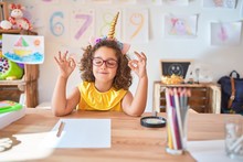 Beautiful Toddler Wearing Glasses And Unicorn Diadem Sitting On Desk At Kindergarten Relax And Smiling With Eyes Closed Doing Meditation Gesture With Fingers. Yoga Concept.