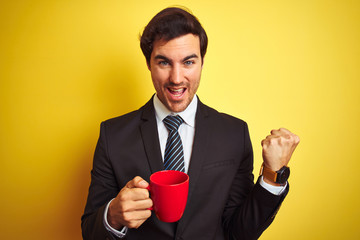 Wall Mural - Young handsome businessman drinking red cup of coffee over isolated yellow background screaming proud and celebrating victory and success very excited, cheering emotion