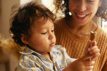 Wall Mural - Close up image of curious little boy with curly hair and chubby face playing with wooden toy sitting on mother's lap. Cropped shot of young Hispanic female smiling broadly, caring for her cute son