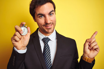 Sticker - Young handsome businessman holding stopwatch standing over isolated yellow background very happy pointing with hand and finger to the side