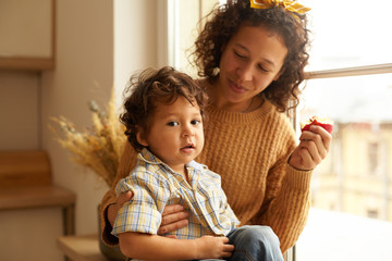 Wall Mural - Indoor shot of joyful young female wearing sweater and headscarf eating apple on windowsill with adorable chubby baby boy on her lap. Family bonds, relationships, love and parenthood concept