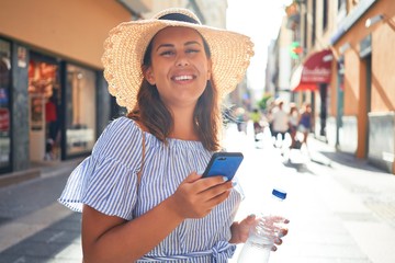 Wall Mural - Young beautiful woman smiling happy walking on city streets on a sunny day of summer using smartphone