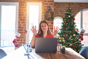 Poster - Beautiful woman sitting at the table working with laptop at home around christmas tree showing and pointing up with fingers number nine while smiling confident and happy.