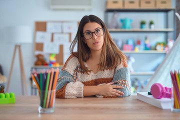 Sticker - Young beautiful teacher woman wearing sweater and glasses sitting on desk at kindergarten Relaxed with serious expression on face. Simple and natural looking at the camera.