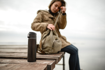 Wall Mural - close-up of a thermos that stands on a village pier against the background of the silhouette of a girl buttoning a backpack