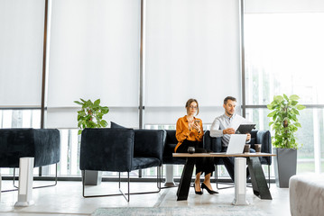 Young couple sitting on the comfortable sofa at the bank or financial institution office, solving some business or legal affairs. Wide view with big window and copy space