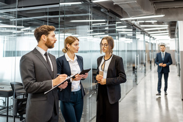 Wall Mural - Group of business people strictly dressed in the suits meeting in the hallway of the modern office building, white-collar workers having informal discussion indoors