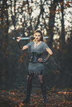 The Girl Knight In Chain Mail With A Metal Sword Cosplay In Gothic Style. Female Warrior Amazon In The Forest Image For Halloween.