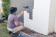 Senior Man Painting The Exterior Of A Brick Suburban Home 
