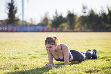 Wall Mural - Young sportive woman in sports clothes training in field at sunrise. Girl standing in plank position on grass in a city park.