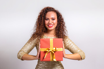 Studio portrait of young woman with dark skin and long curly hair wearing tight sexy dress, holding wrapped present with golden bow. Close up, copy space for text, isolated background.