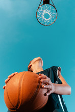 Sports And Basketball. A Young Teenager In A Black Tracksuit Stands With A Ball In His Hands And Prepares To Throw The Ball Into The Ring. Blue Sky With Hoop. Bottom View. Copy Space