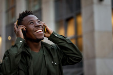 Happy African-american Man In Stylish Jacket In Wireless Headphones Listening Music On The Street Of The Evening City. Space For Text