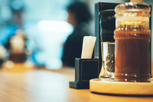 Closeup, Black Pepper And Dipping Sauce In Glass Bottles On Round Wooden Condiment Tray, Toothpicks In White Package And Napkins In Leather Box On Table. Blurred Waitress And Chef Talk In Background.