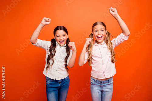 Portrait of ecstatic two children girl win competition feel crazy scream yeah raise fists wear modern outfit isolated over orange color background