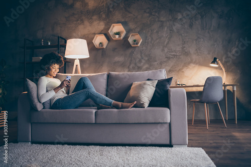 Profile photo of pretty dark skin curly lady domestic atmosphere texting telephone lying comfy couch in lamp light wear casual outfit evening living room indoors
