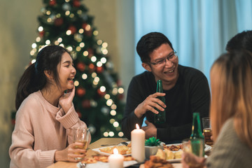 Merry Christmas and Happy New Year! Young friends group having dinner at home. Asian family party with beer and pizza food near christmas tree at house in night. Celebration holiday and togetherness.