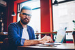 © BullRun - Portrait of bearded hipster guy in optical spectacles looking at camera during break from working remotely on modern netbook in cafeteria, handsome young man dressed in casual wear sitting with laptop