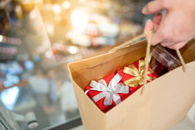 Male Hand Holding Paper Shopping Bag With Red Gift Boxes Inside At Christmas Event In Department Store. Buying Present For Xmas Holiday And New Year Celebration Concept