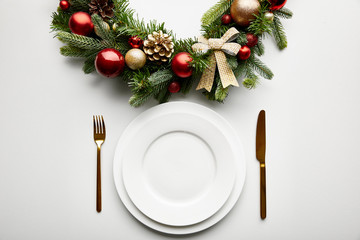 Wall Mural - top view of white plates with golden cutlery near festive christmas wreath with baubles on white bac