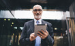 © BullRun - Confident senior businessman in suit and eyeglasses surfing tablet in front of commercial building on street