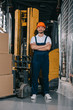 © LIGHTFIELD STUDIOS - handsome warehouse worker smiling at camera while standing near forklift loader with crossed arms
