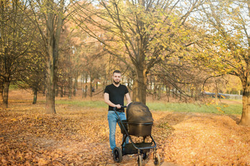 A young man with a beard walks in the park with a black pram