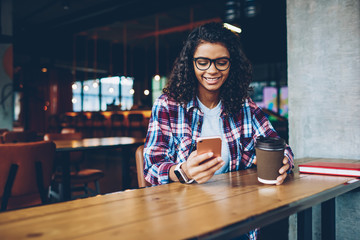 Cheerful african american female blogger with curly hair laughing during online chat with friends on smartphone connected to free wifi.Positive teen publishing funny story on cellular sitting indoors