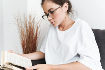 Sticker - Image of serious young woman reading book while sitting on sofa