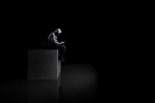  A Lonely Man With Smartphone Sits On A Concrete Cube In The Dark. Reflection On The Floor.