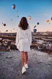 Fototapeta Koty - A tourist girl wearing white sweater on a mountain top enjoying wonderful view of balloons in Cappadocia