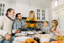 The Family Sits Down For Dinner On Thanksgiving. Young Woman Serves A Festive Turkey With A Salad, Grandfather, Father And Children Sit And Look At The Tasty Food And Smile