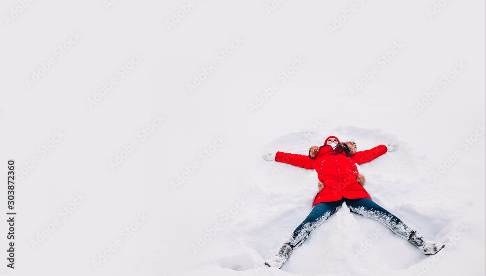 Happy young woman in red lying on snow and making snow angel figure with hands and legs - obrazy, fototapety, plakaty 