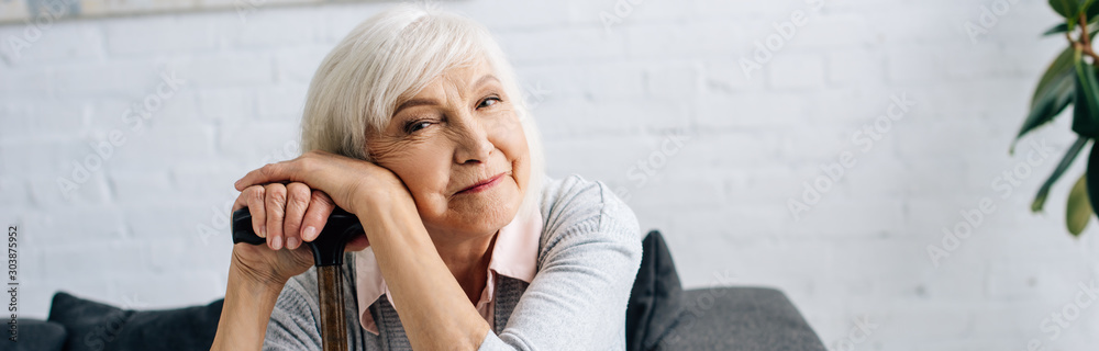 panoramic shot of smiling senior woman with wooden cane looking at camera in apartment - obrazy, fototapety, plakaty 