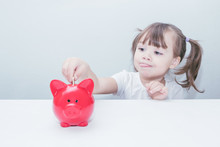 Child Girl Dips A Coin In A Red Piggy Bank Standing On The Table