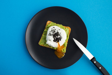 Wall Mural - Sandwich with poached egg and avocado on the black plate in the center of the blue background. Top view. Copy space.