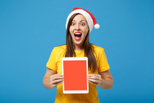 Excited Woman Santa Girl In Yellow T-shirt Christmas Hat Posing Isolated On Blue Background. New Year 2020 Celebration Holiday Concept. Mock Up Copy Space. Hold Tablet Pc Computer With Blank Screen.