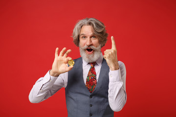 Amazed elderly gray-haired mustache bearded man in classic shirt vest tie isolated on red background. People lifestyle concept. Mock up copy space. Hold bitcoin, future currency point index finger up.