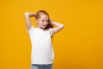 Beautiful little ginger kid girl 12-13 years old wearing white t-shirt isolated on yellow background children portrait. Childhood lifestyle concept. Mock up copy space. Holding hair, looking aside.