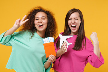 Wall Mural - Two women friends european african girls in pink green clothes posing isolated on yellow background. People lifestyle concept. Hold passport ticket airplane showing victory sign doing winner gesture.