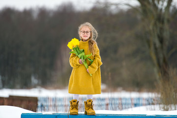 Adorabe little girl wearing adult oversized sweater and big fathers boots. Beautiful young  girl in glasses standing on bench with bouquet of yellow tulips in snowy winter day.