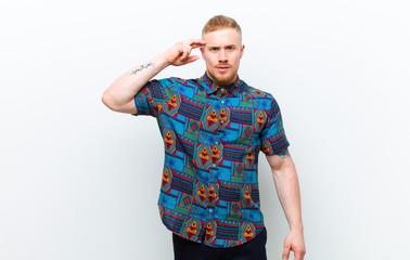 young blonde man wearing a cool shirt feeling confused and puzzled, showing you are insane, crazy or out of your mind against white background