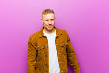 young blonde man looking happy and friendly, smiling and winking an eye at you with a positive attitude against purple background