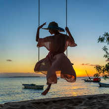 Beautiful Girl In A Straw Hat And Pareo Swinging On A Swing On The Beach During Sunset Of Zanzibar Island, Tanzania, Africa. Travel And Vacation Concept
