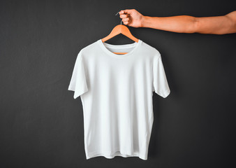close up of hand holding white color t-shirt hanging on wooden cloth hanger over black color background, copy space