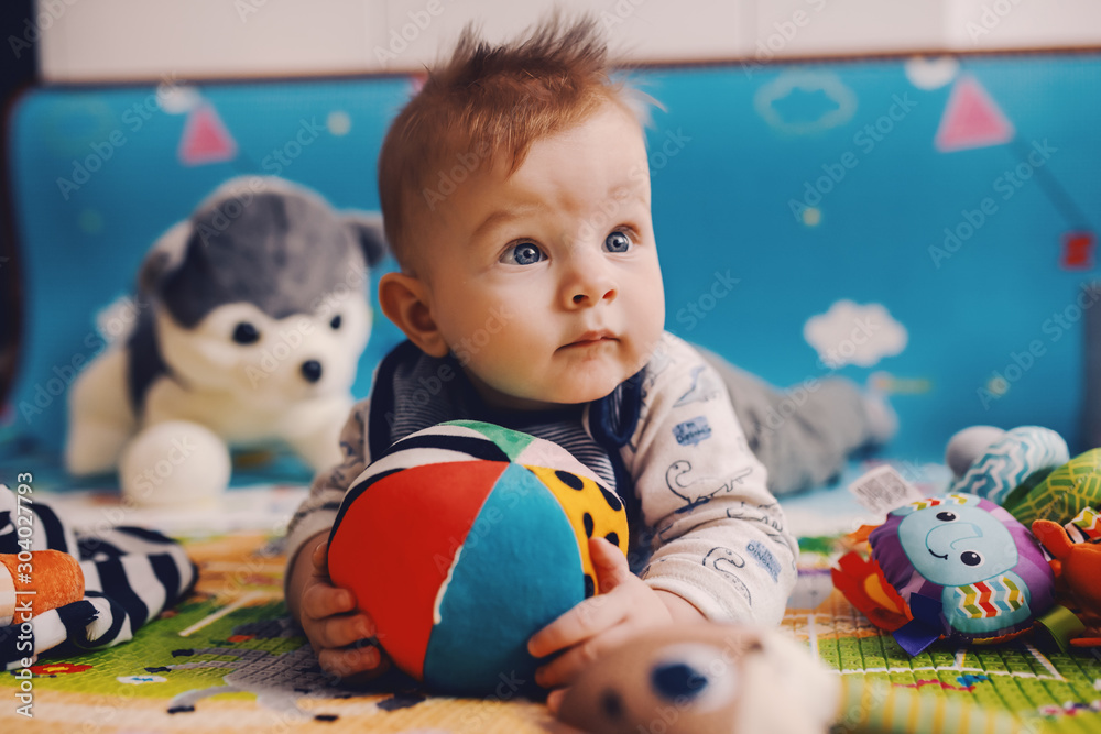 Adorable curious caucasian baby boy with big beautiful blue eyes lying on the floor on stomach, holding stuffed ball and looking away. - obrazy, fototapety, plakaty 